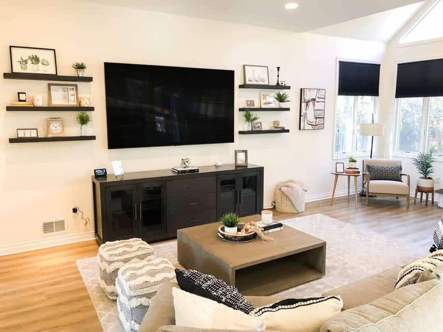 Modern living room with a large TV, black wall shelves, a cozy sofa, patterned poufs, and decor accents beautifully illuminated by bright natural light from windows