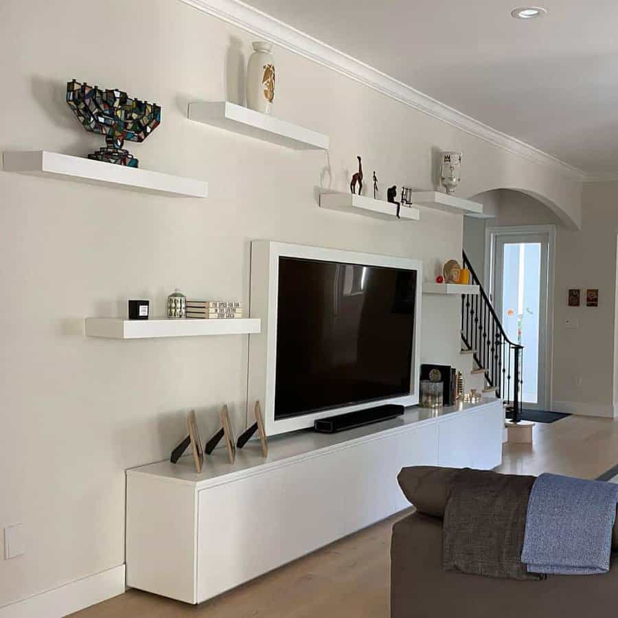 Modern living room with a large TV, white shelves displaying decor items, and minimalistic furniture featuring light wood flooring