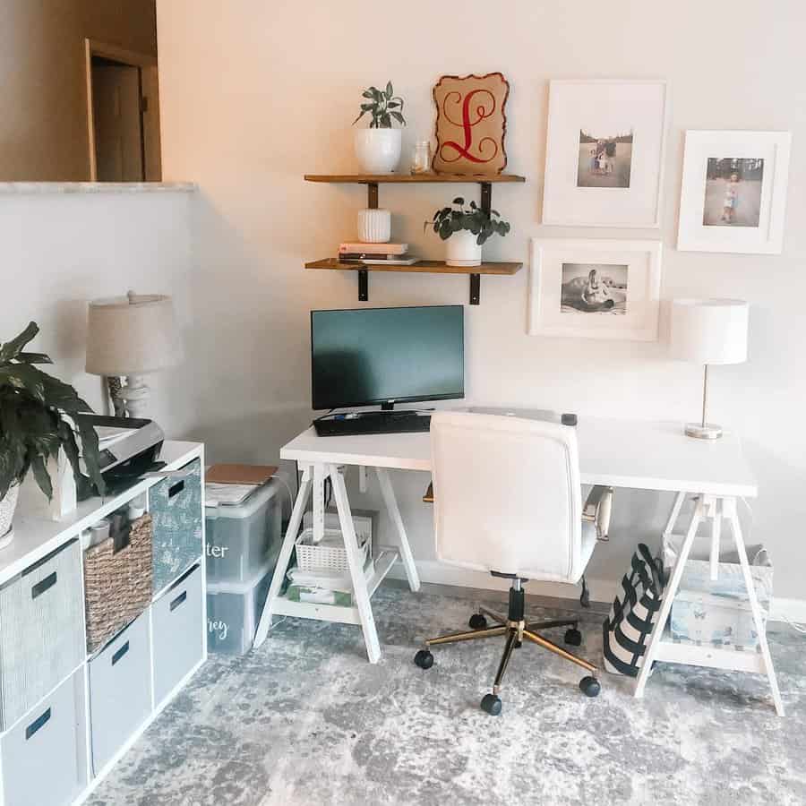 Home office with a white desk, chair, computer, and lamps; shelves with decor above, framed photos on the wall, and storage cubes nearby