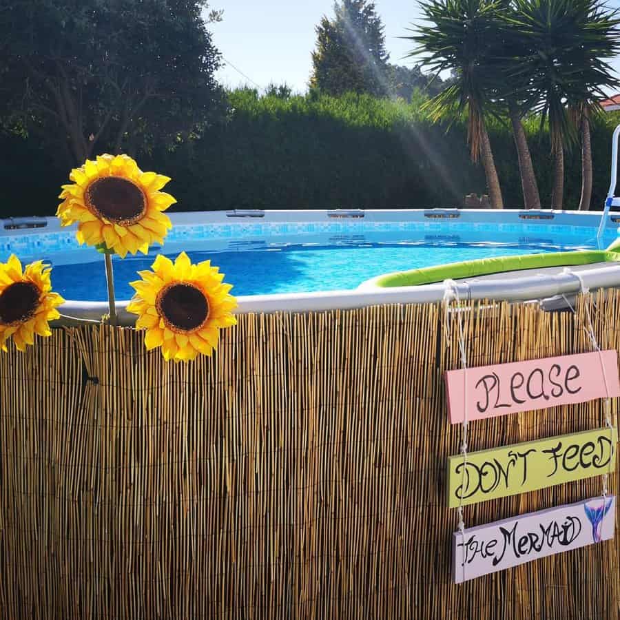 Above-ground pool with a straw fence, sunflowers, and sign saying "Please Don't Feed the Mermaids" with trees in the background