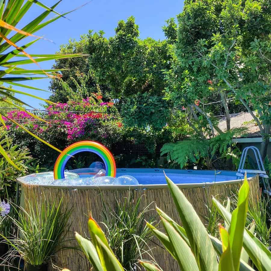 Above-ground pool with a rainbow float surrounded by lush greenery and flowers under a clear blue sky