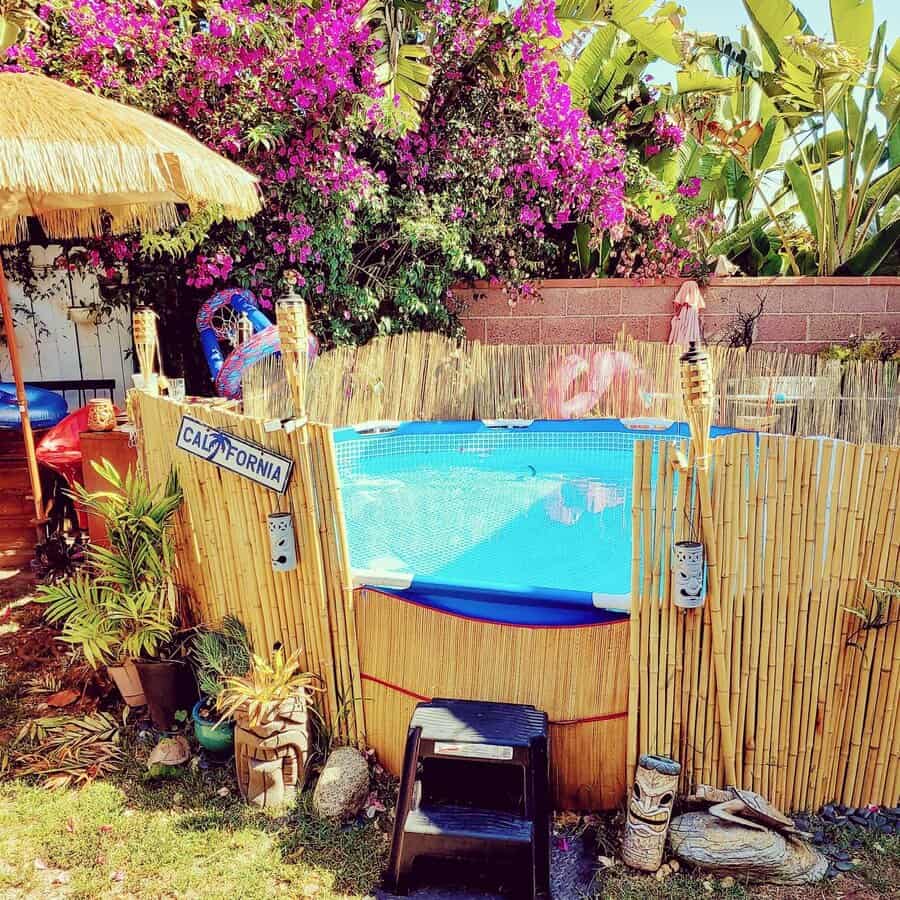 Round above-ground pool with bamboo fencing, surrounded by vibrant flowers and tropical plants, a "California" sign adds a playful touch