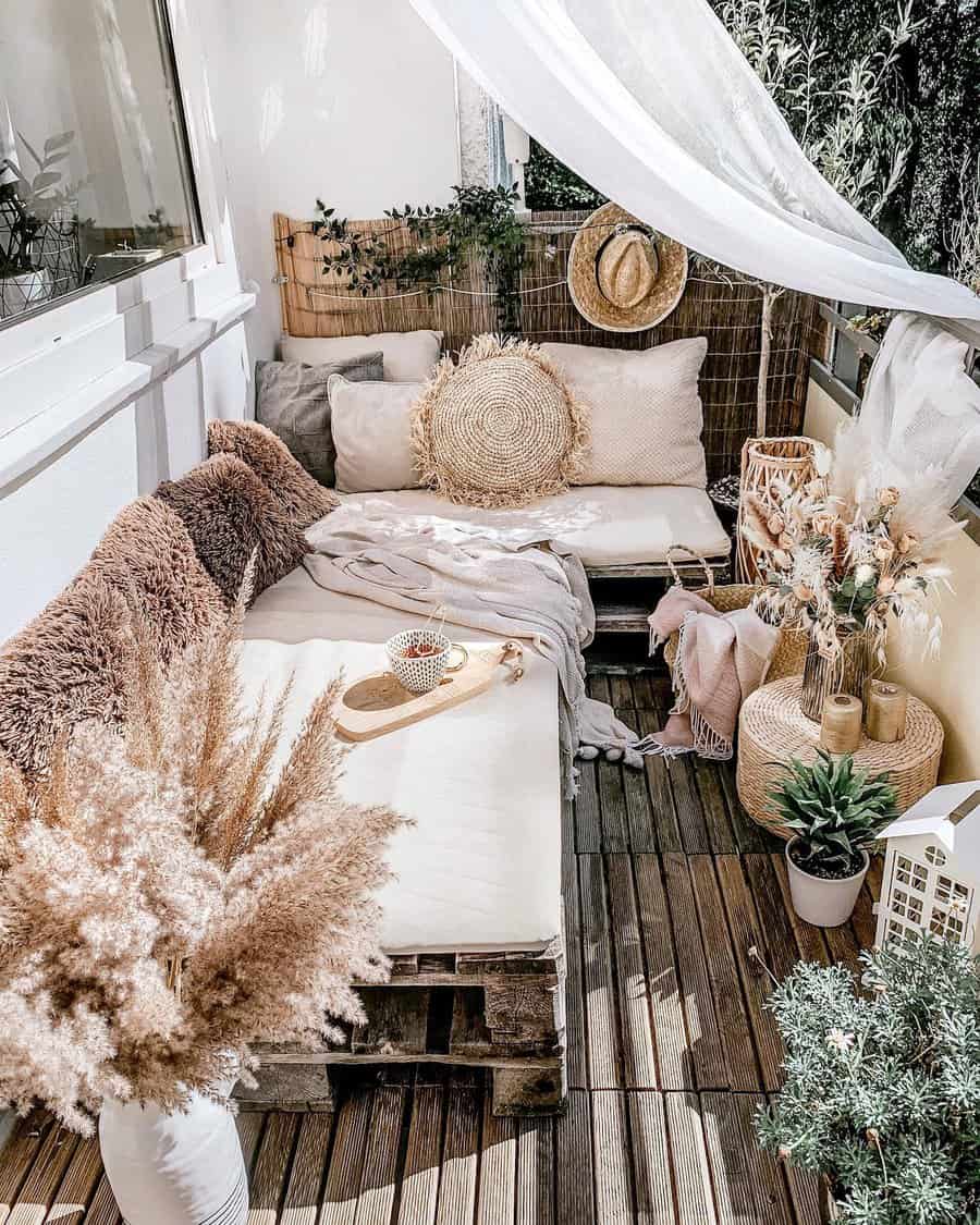 Boho-style small balcony with a cozy pallet sofa, neutral cushions, and soft blankets. A sheer white curtain adds an airy touch, while pampas grass, woven decor, and potted plants enhance the natural aesthetic.