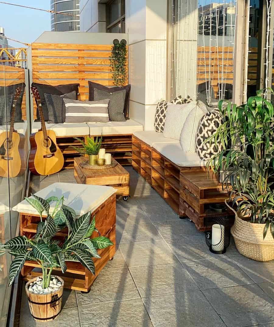 Modern small balcony with DIY wooden pallet seating, neutral cushions, and black-and-white accent pillows. Two guitars rest against a glass railing, while potted plants and string lights add warmth to the space.