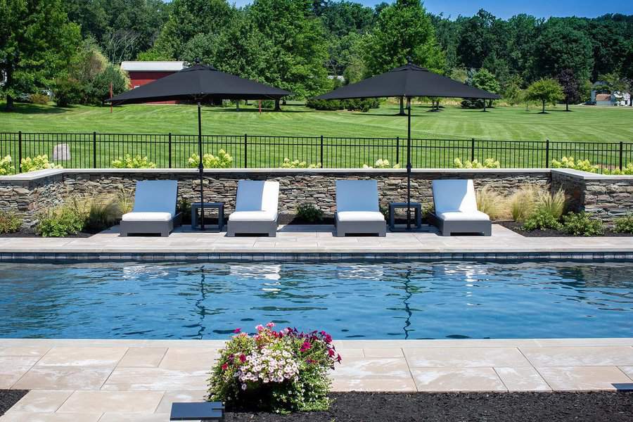 Outdoor swimming pool with four white lounge chairs and umbrellas by a stone wall, overlooking a grassy field and trees