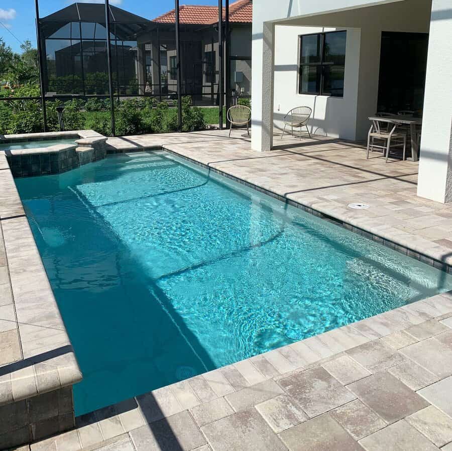 Rectangular outdoor pool with clear blue water, surrounded by a tiled patio. Nearby are chairs and a table under a covered area