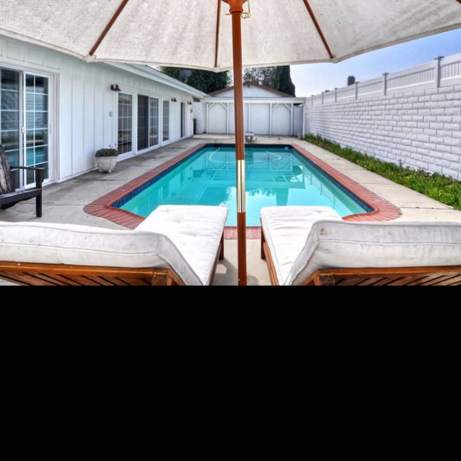 Backyard with a white house, swimming pool, and cushioned seating under an umbrella, bordered by a white fence and plants