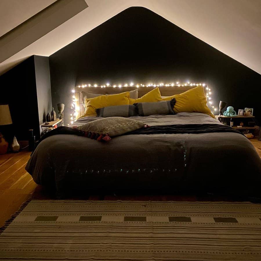 Cozy bedroom with a string-lit headboard, yellow and gray bedding, and a rug on wooden floor; sloped black ceiling adds charm