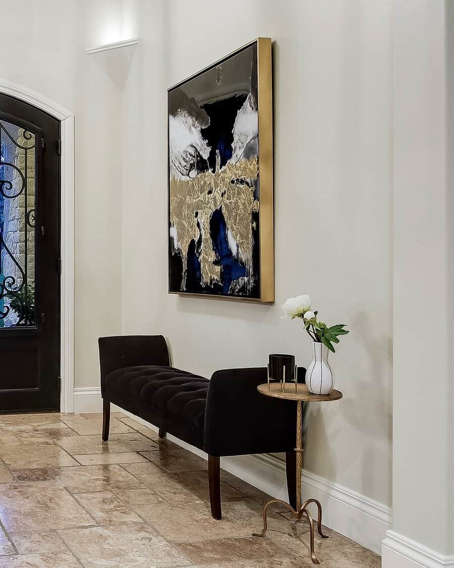 Hallway with a black bench, abstract painting, and small table holding a white vase with a single flower near a black ornate door