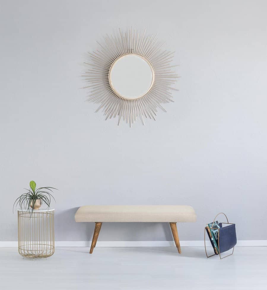 Minimalist room with a sunburst mirror, light bench, potted plant on a wire table, and a magazine holder on a light gray wall