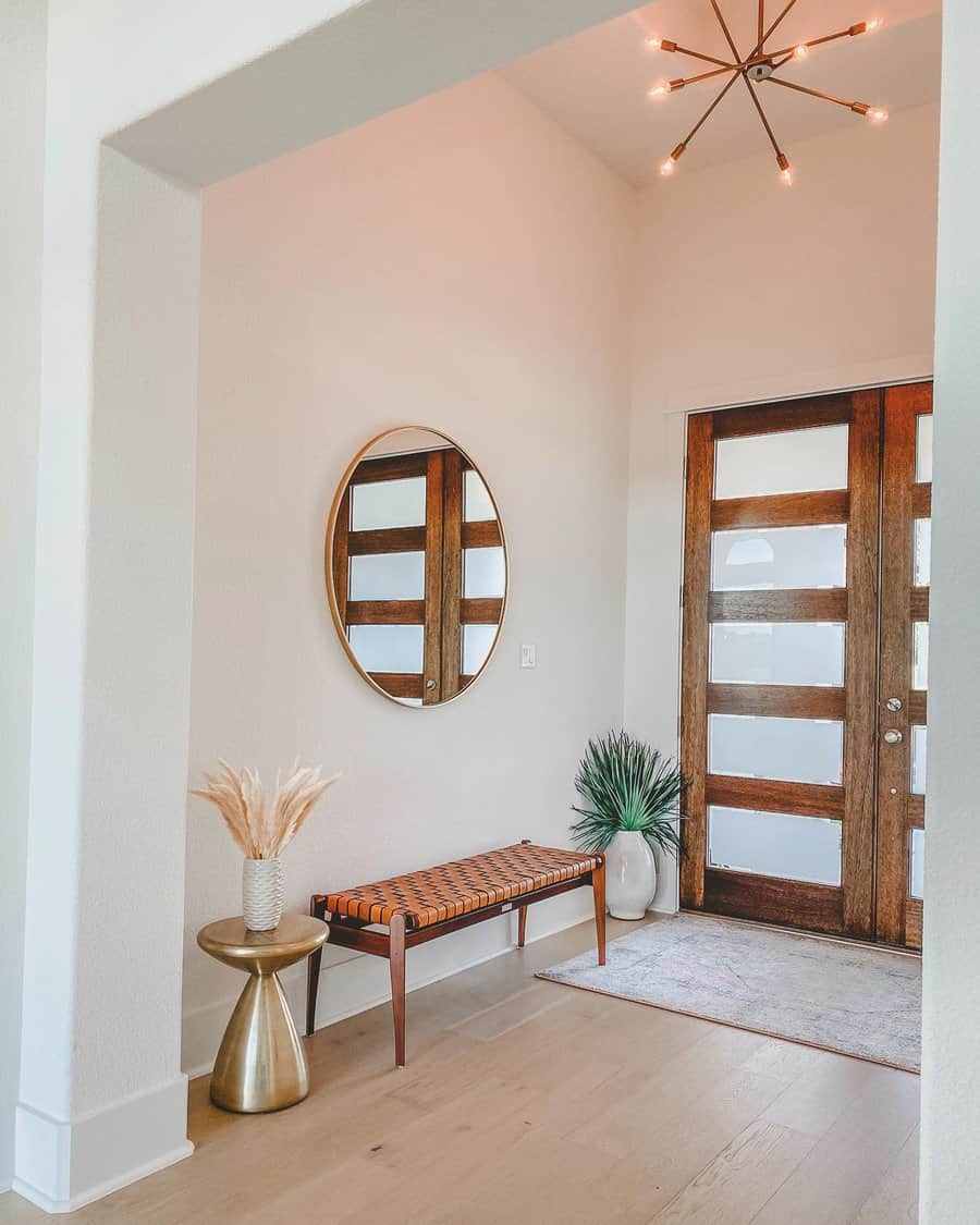 Modern entryway with wooden door, round mirror, starburst light, plant, vase with dried grasses, and patterned bench on light wood floor