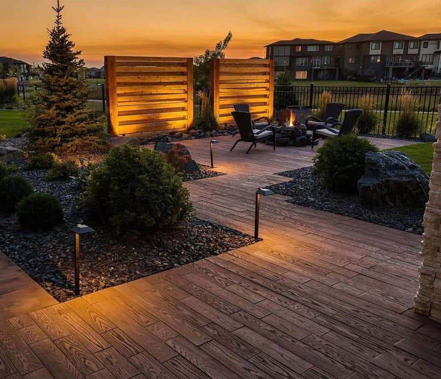 Luxurious backyard with a wooden deck, illuminated privacy panels, fire pit area, lush greenery, and a scenic sunset view