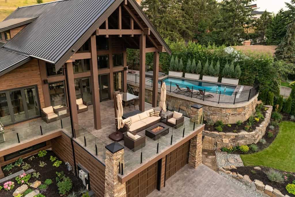 Luxury outdoor space featuring an elevated patio with modern seating, a fire pit, a stone-lined pool area, and lush landscaping