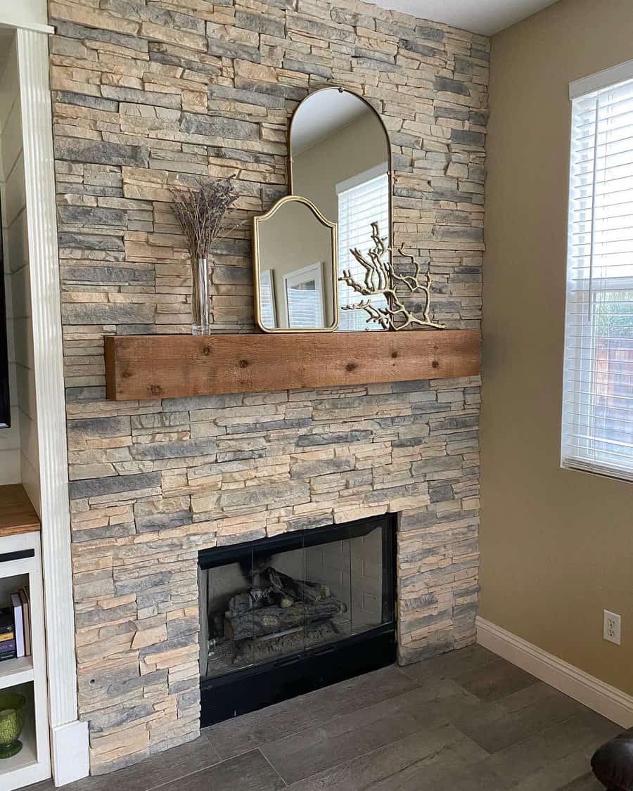 Stone fireplace with a wooden mantel featuring decorative mirrors, dried branches, and a coral piece by a window with blinds