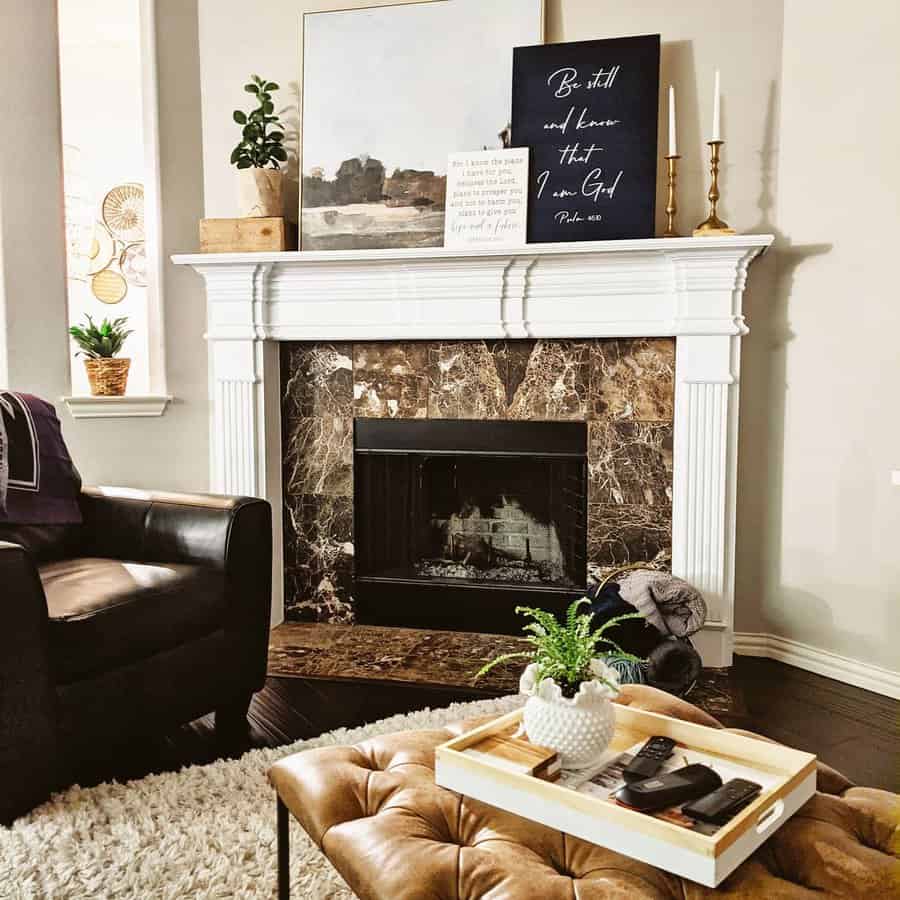 Cozy living room with a fireplace, artwork on the mantel, a brown leather ottoman, and a tray with a plant and remote controls