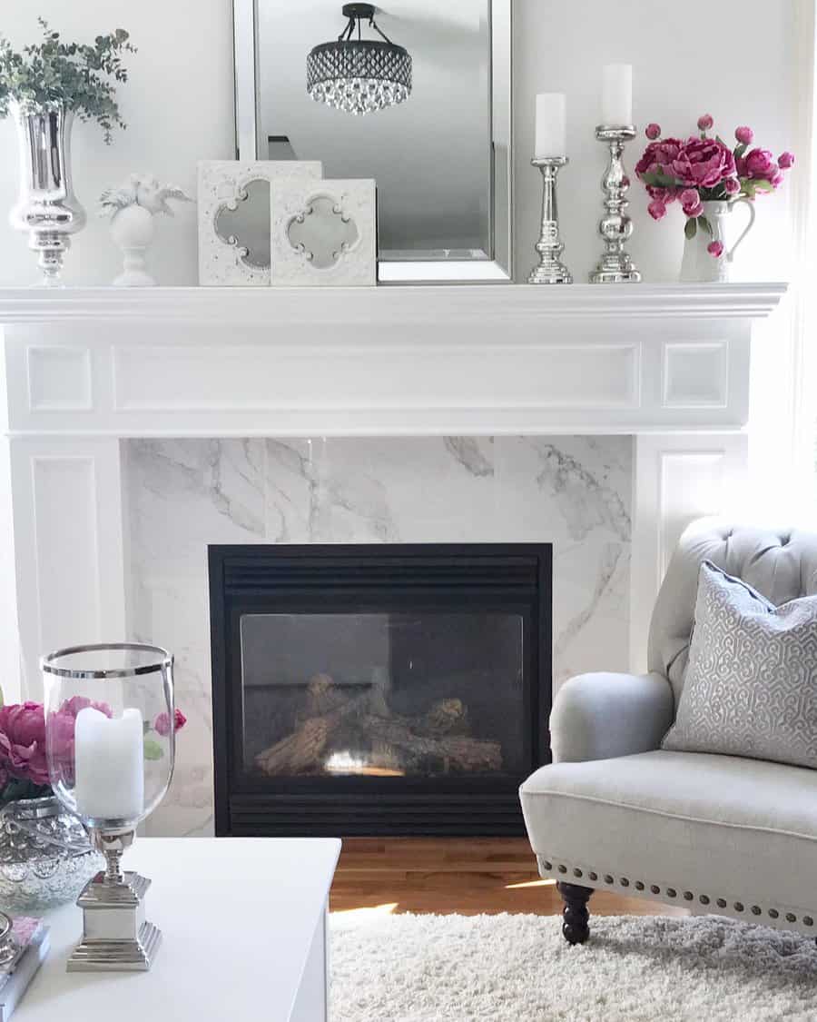 A cozy living room with a marble fireplace, decorated mantel, gray armchair, and pink flowers, creating a chic and elegant ambiance