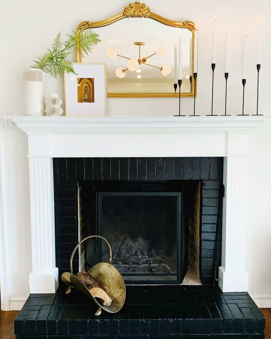 Black fireplace with white mantel, gold mirror, candles, art, and a vase; a log holder sits on the hearth