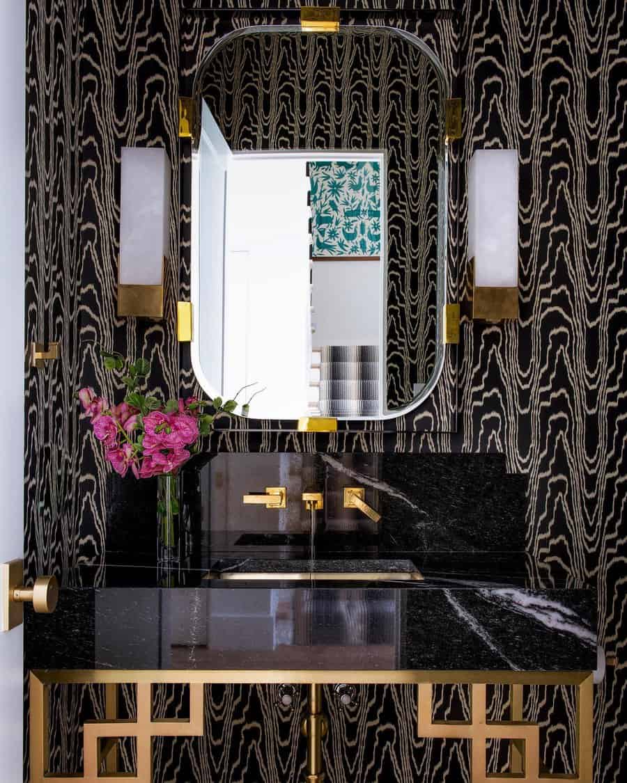 Glamorous bathroom with black marble backsplash and countertop, gold fixtures, bold patterned wallpaper, and a statement mirror.