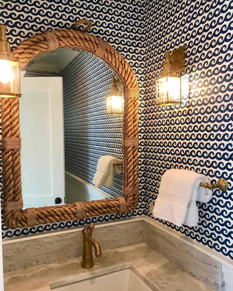 Coastal-style bathroom with beige marble backsplash, rope-framed mirror, brass fixtures, and navy wave-patterned wallpaper.