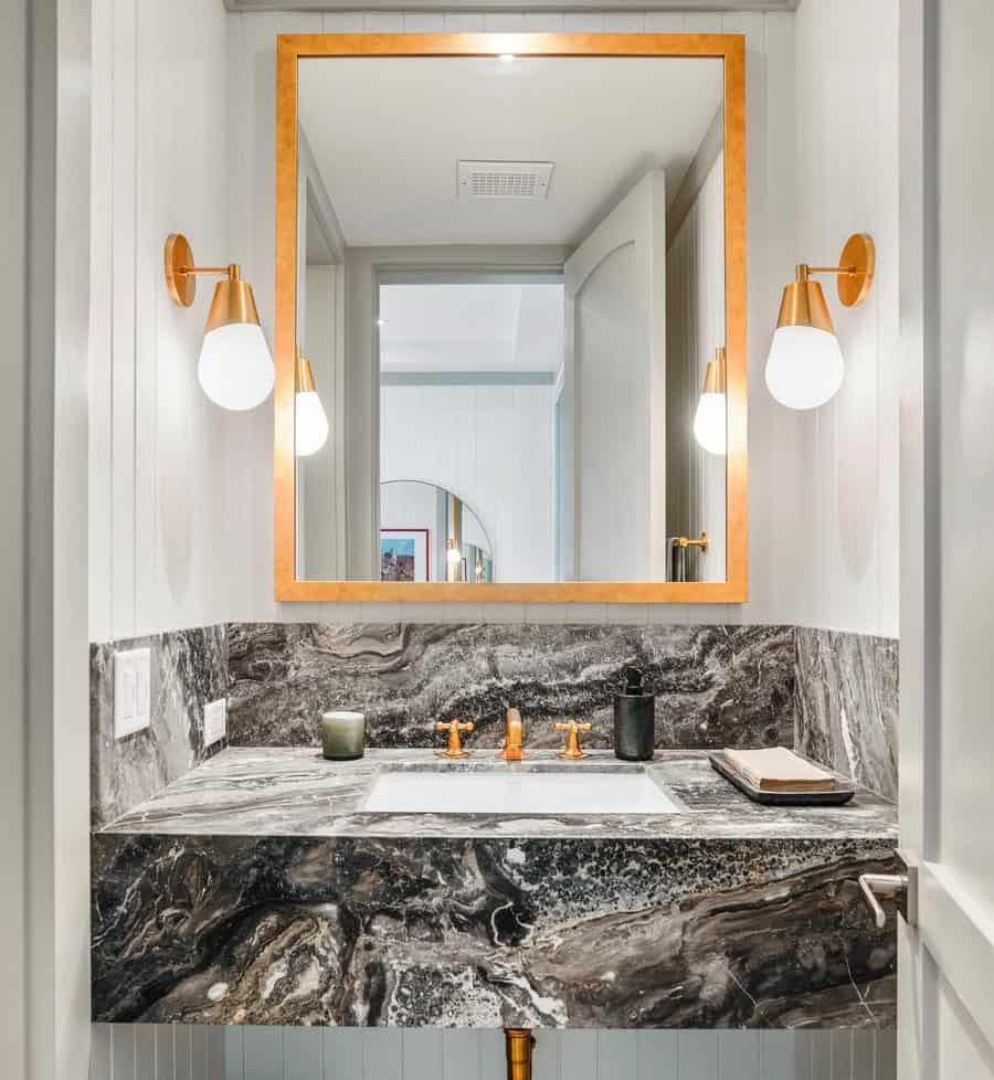 Bold bathroom with swirling black marble vanity and backsplash, gold fixtures, wooden-framed mirror, and modern wall sconces.