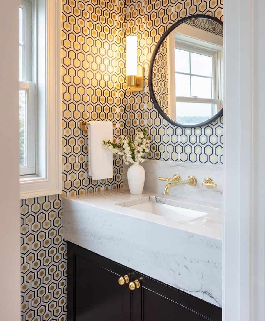 Chic bathroom with bold honeycomb wallpaper, white marble backsplash and sink, gold fixtures, and a round black-framed mirror.