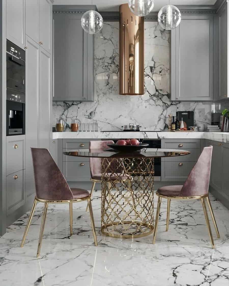 Luxurious kitchen with marble floors and backsplash, gold accents, round table, gray cabinets, and modern chairs under glass pendant lights