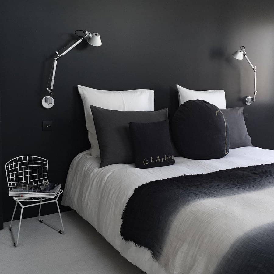 Minimalist bedroom with dark walls, white bedding, gray pillows, wall-mounted lamps, and a small metal chair with magazines