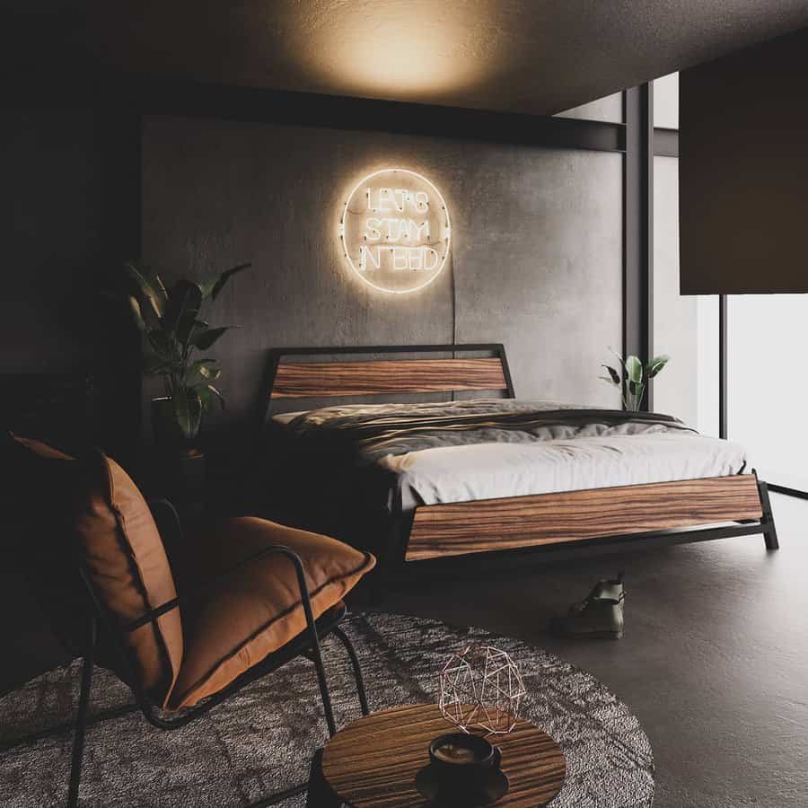 Industrial bedroom with a wooden bed, brown chair, and a neon sign reading "Let's Stay In Bed" on a dark wall, surrounded by potted plants