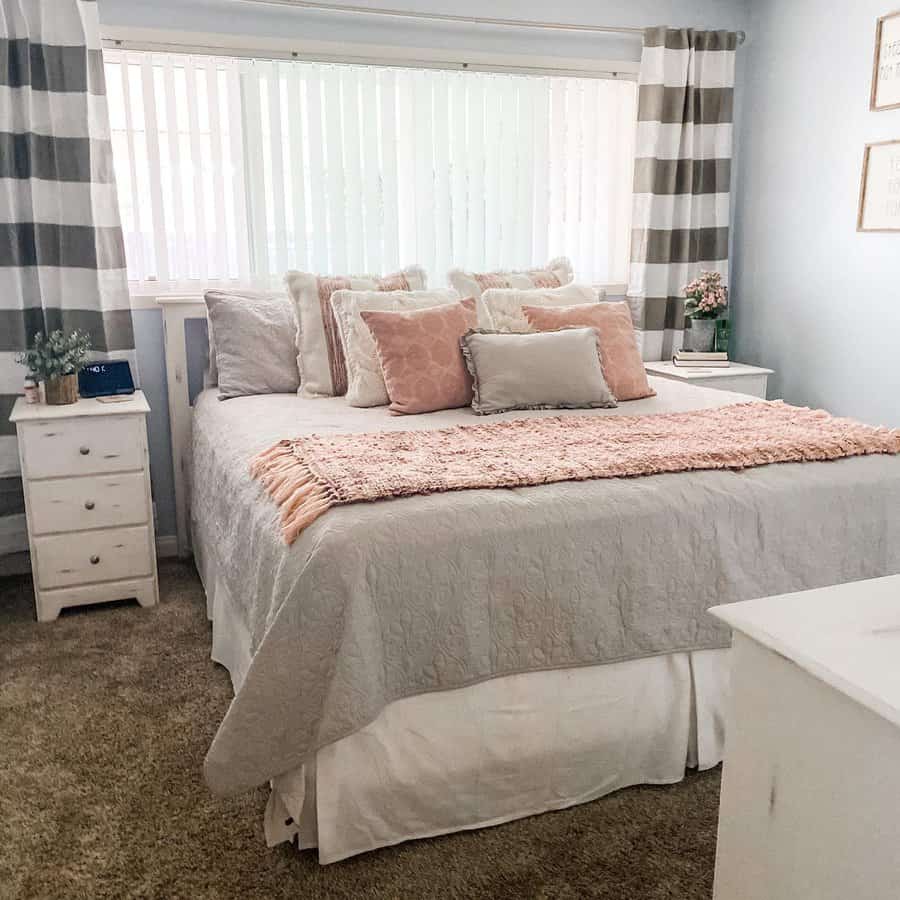 Cozy bedroom with a bed covered in white and pink bedding, striped curtains, bedside tables, and soft lighting through blinds