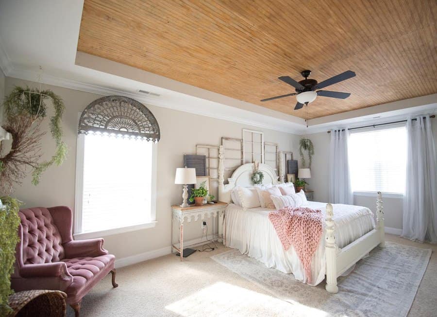 Bright, cozy bedroom with a white bed, wooden ceiling, ceiling fan, large window with sheer curtains, plants, and a pink armchair