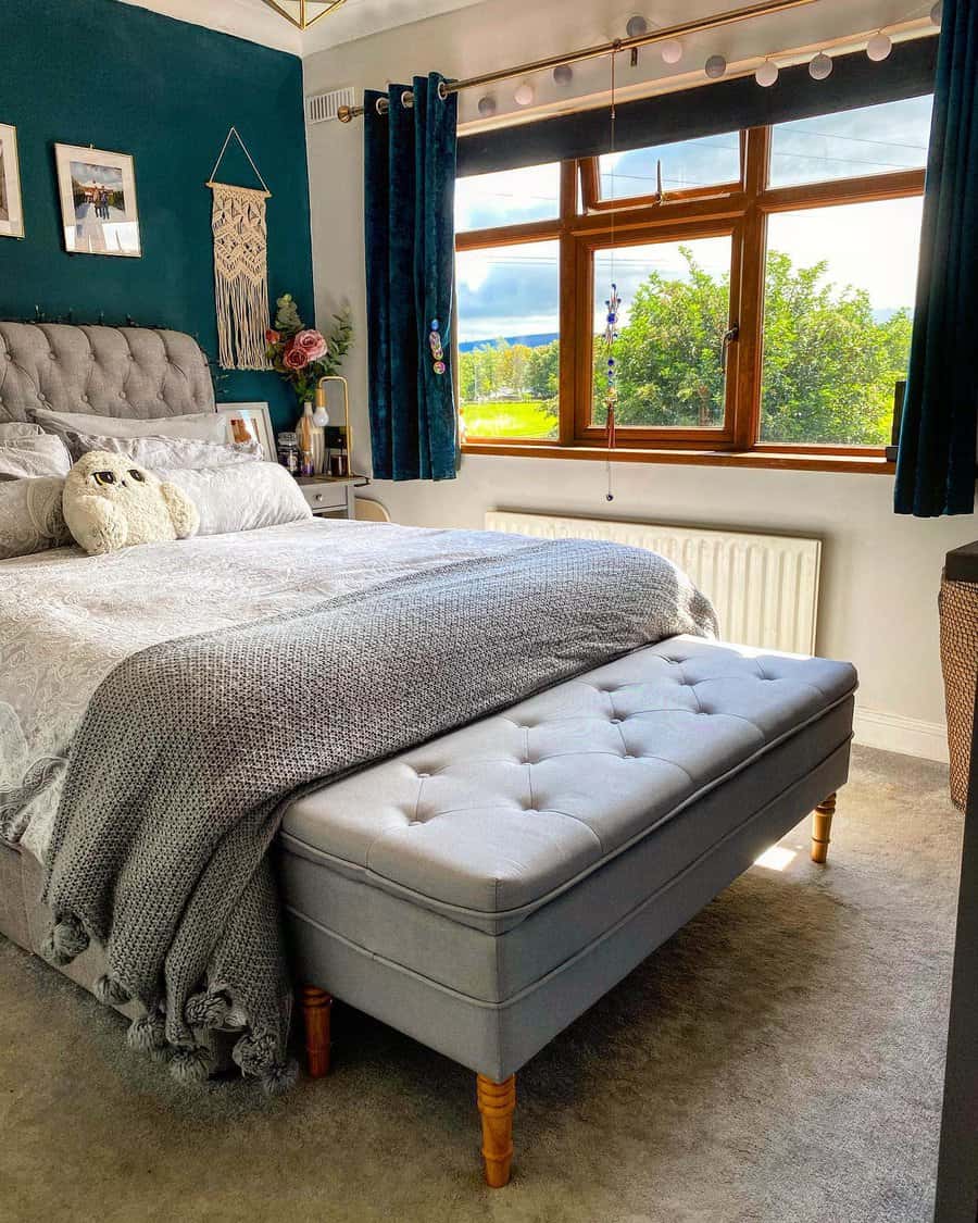 Cozy bedroom with teal walls, tufted headboard, gray bedding, a bench at the foot of the bed, and a window view of greenery