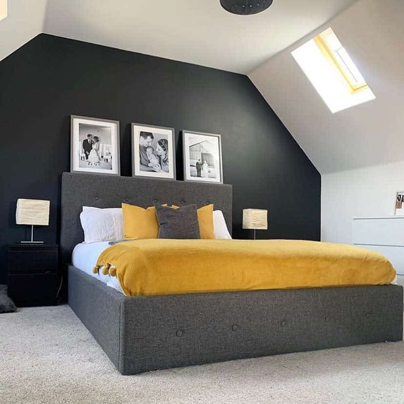 Modern bedroom with a gray bed, mustard blanket, black accent wall, and skylight; black-and-white photos hang above the bed