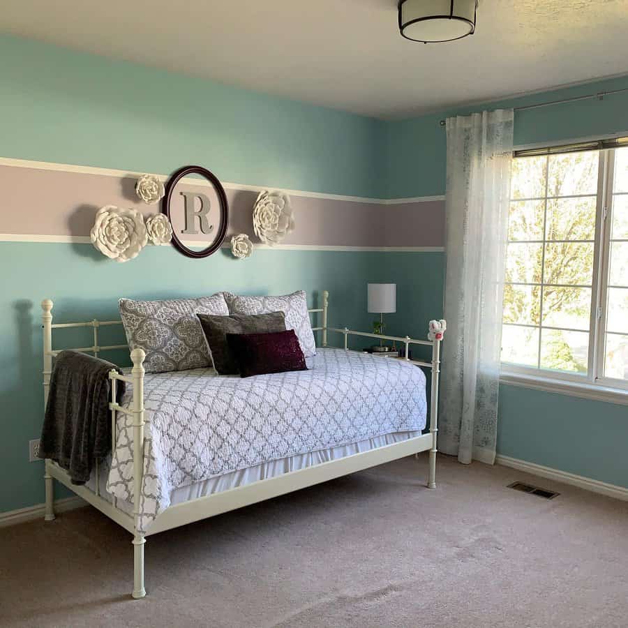 Light blue bedroom with a white metal frame daybed, floral decor, pillows, and a window with sheer curtains featuring a decorative "R" hanging above the bed