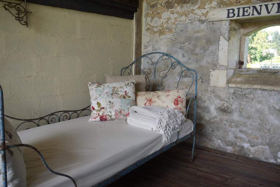 Antique metal daybed with floral pillows and a knit blanket, set against a rustic stone wall with a "BIENVENUE" sign