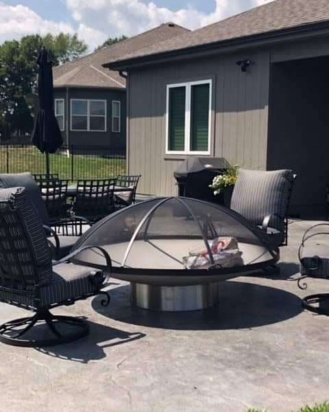 Outdoor patio with a round metal fire pit, covered by a mesh dome, surrounded by striped cushioned chairs and a grill nearby