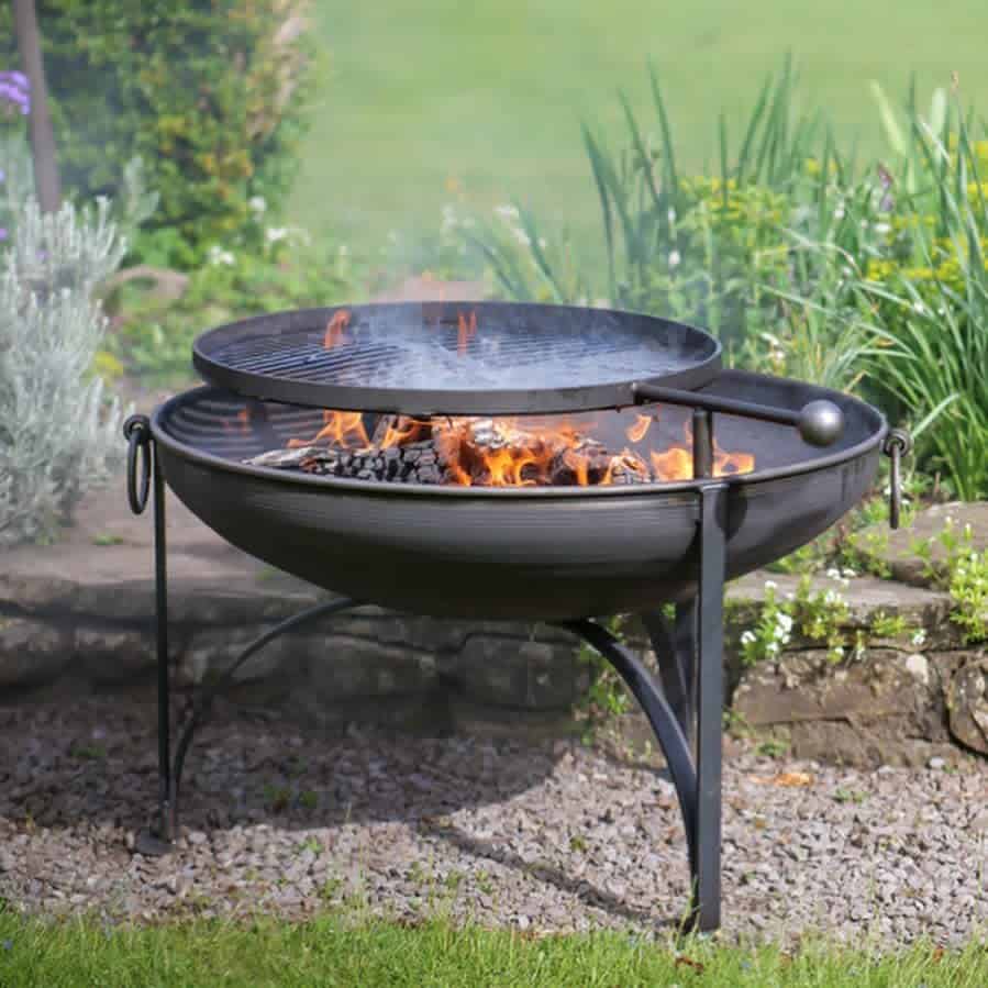 A black metal fire pit with burning logs and a round grill, set on grass with greenery in the background