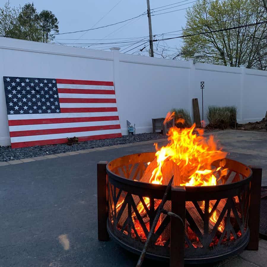 Fire pit lit on patio with large American flag panel on white fence, surrounded by grass and trees