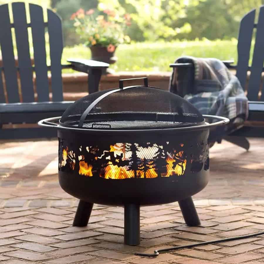 A black metal fire pit with a flame design sits on a brick patio, surrounded by Adirondack chairs and a plaid blanket draped over one chair