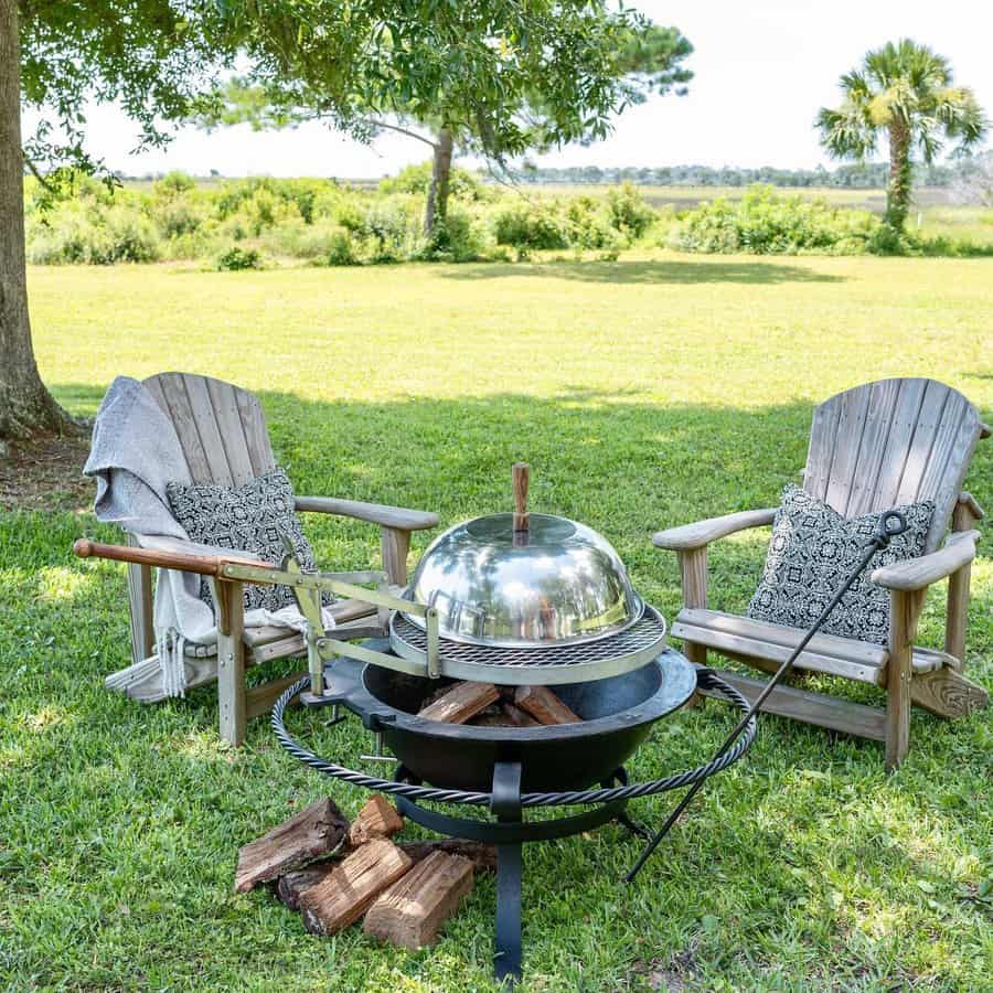 Two wooden chairs with cushions sit around a steel fire pit on a grassy lawn, with trees and greenery in the background