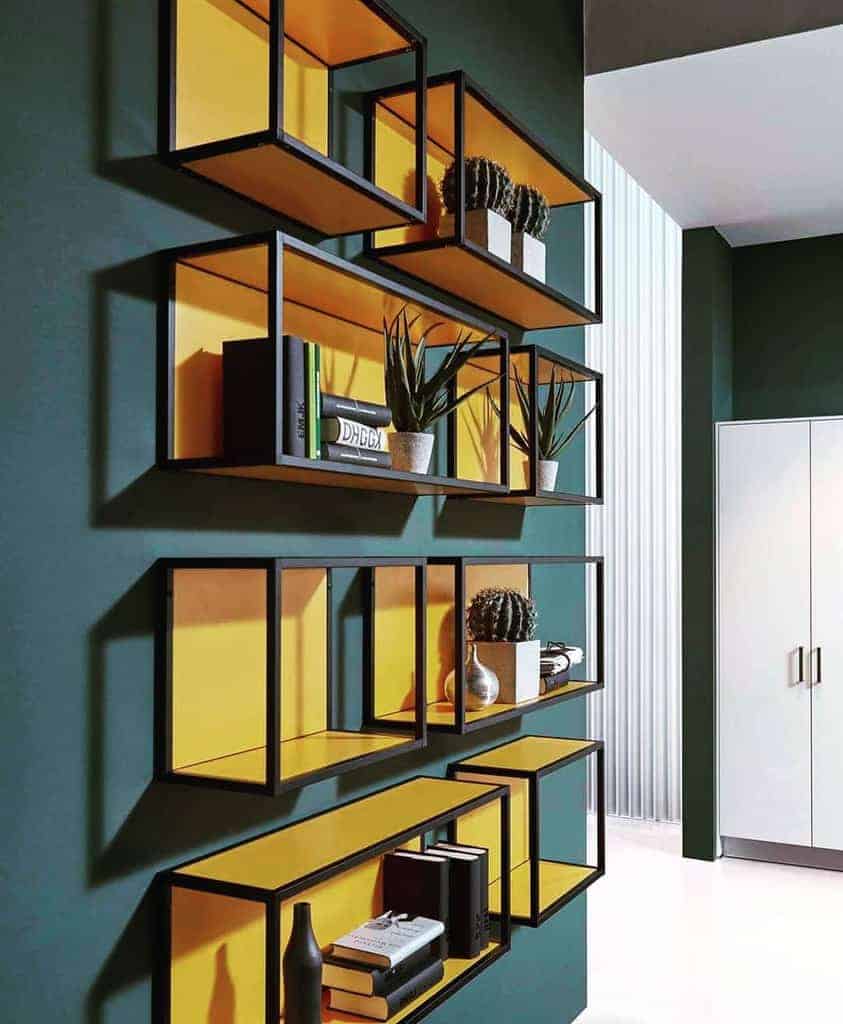 Geometric wall shelves with books, cacti, and a vase on a dark green wall, bright yellow interiors add contrast