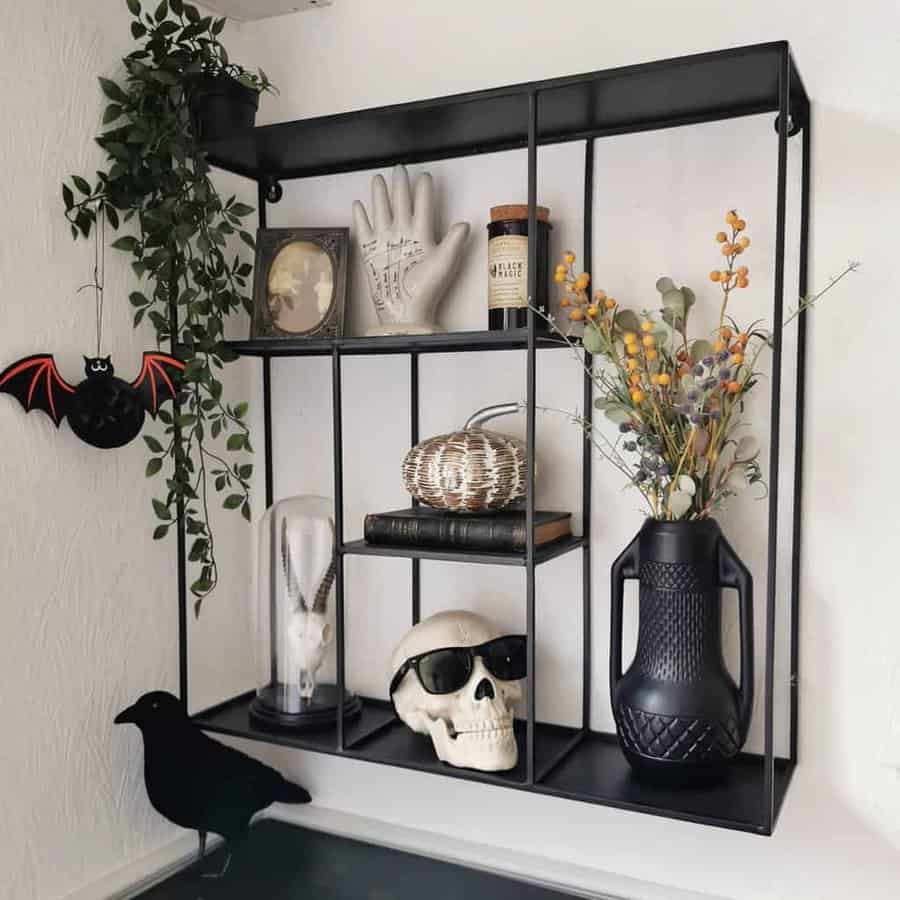 A black wall shelf with a skull, decorative bat, crow, vase, and various curiosities, including plants and trinkets, arranged on it