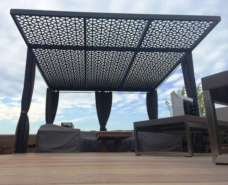 Outdoor patio with a metal pergola and draped curtains, featuring lounge chairs and a wooden floor under a partly cloudy sky