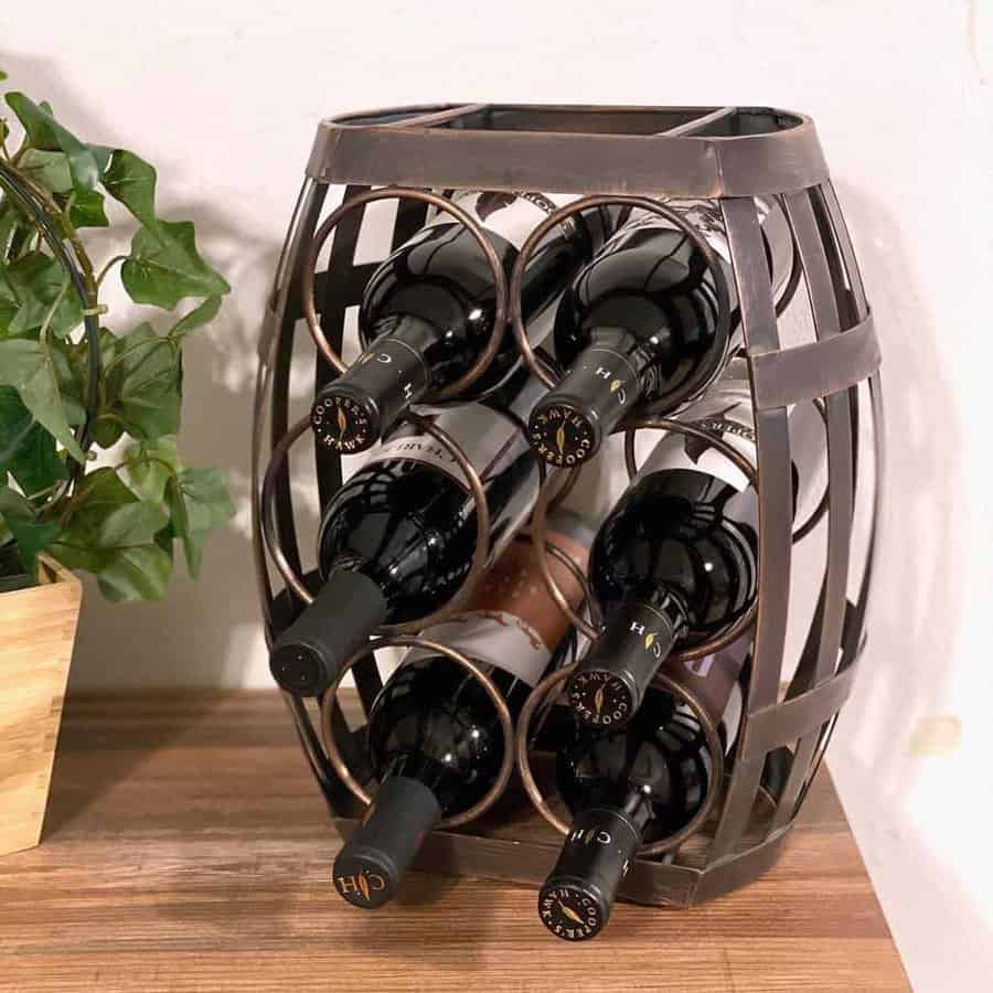 Wine rack shaped like a barrel, holding six bottles, on a wooden surface next to a potted plant