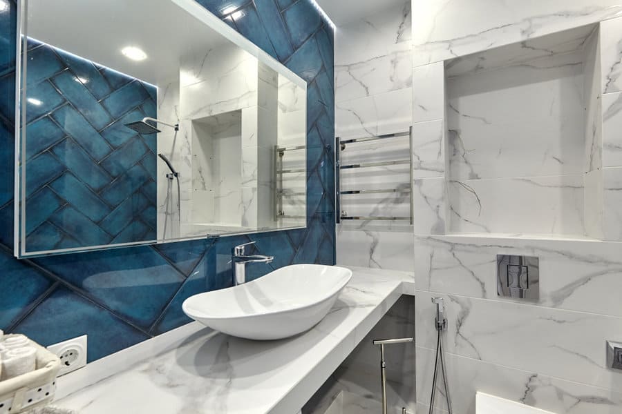 Modern bathroom with marble walls, blue accent tiles, and a vessel sink on a white countertop. Large mirror and towel rack visible