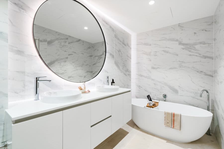 Modern bathroom with large circular mirror, white vanity with dual sinks, freestanding bathtub, and marble-tiled walls