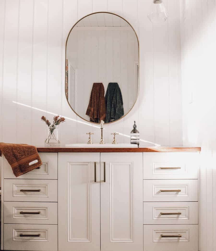 A bathroom with a white vanity, oval mirror, and gold faucets, with towels hanging behind and a brown towel draped on the counter