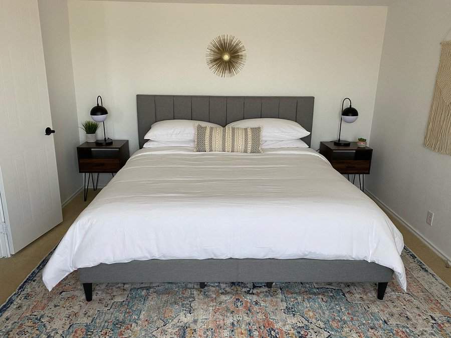 A neatly made bed with gray headboard, white linens, and two pillows, nightstands with lamps on either side and a sunburst wall decor