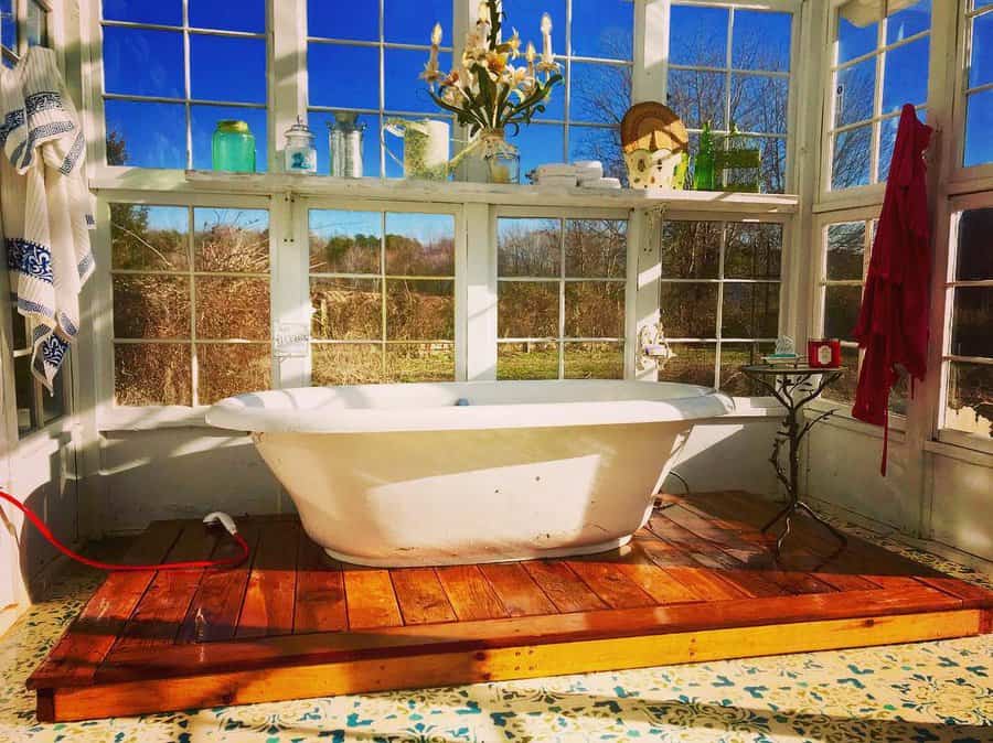 Sunlit bathtub in a glass-enclosed space with panoramic countryside views, blending rustic charm with cozy farmhouse aesthetics