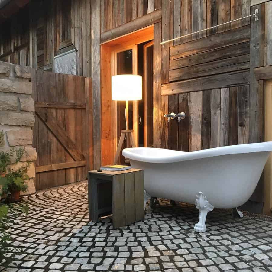 Outdoor clawfoot bathtub in a rustic setting with cobblestone floor, wooden walls, and a glowing floor lamp beside a small table