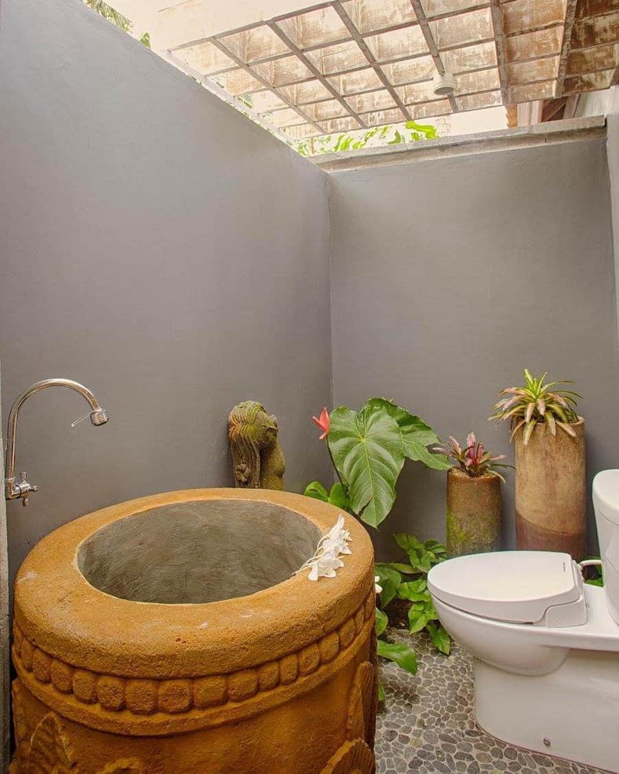 Outdoor bathroom with a stone tub, white toilet, plants, and gray walls under a transparent ceiling panel